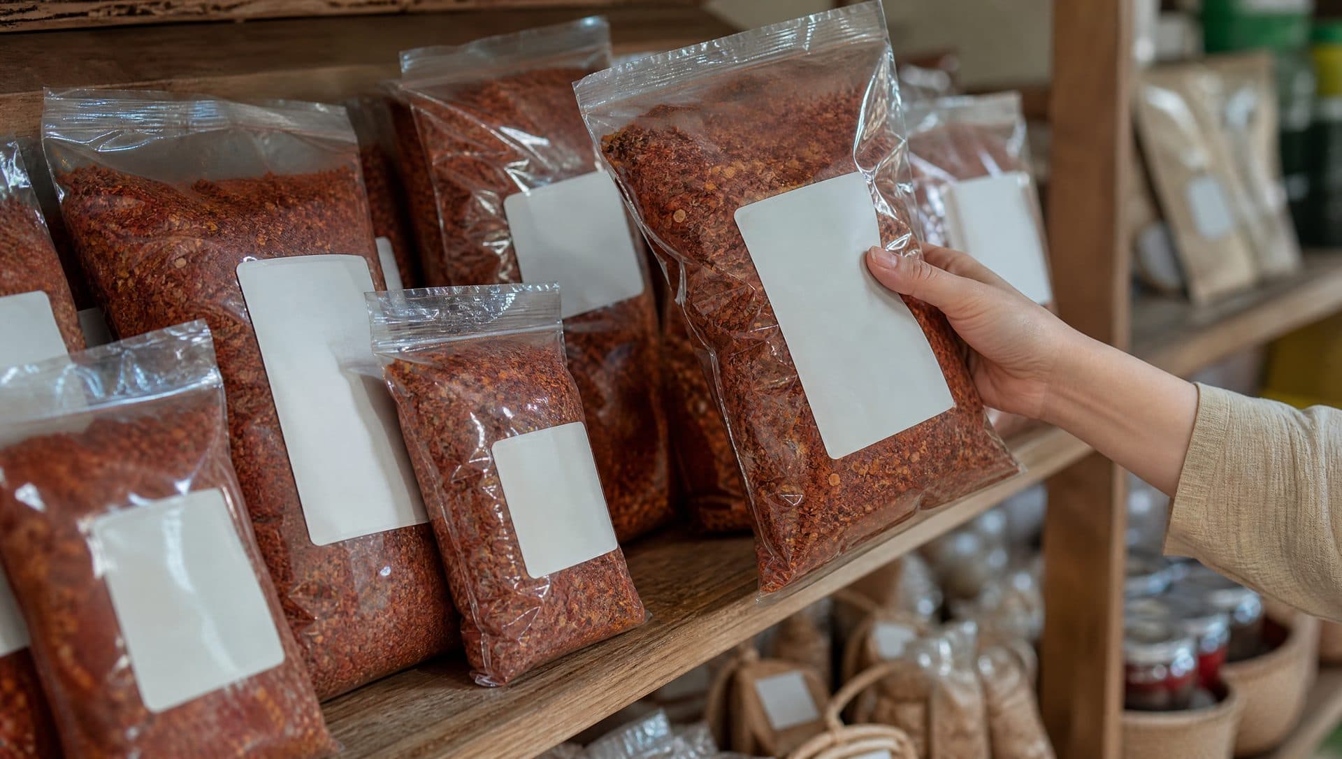 A hand lifting a clear bag of coarse red Korean chilli flakes from a wooden grocery shelf lined with more bags of the same flakes in several sizes