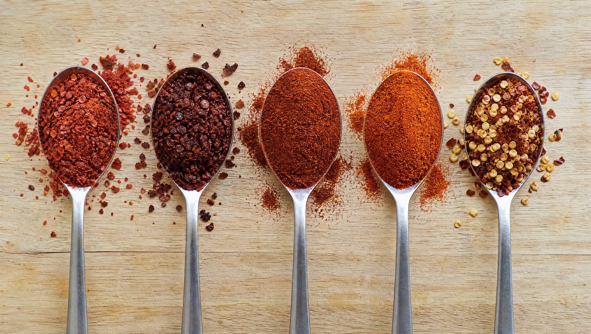 Five spoons in a row on a wooden board holding coarse red Korean chilli flakes, dark oily Aleppo pepper flakes, fine paprika, fine cayenne and crushed red pepper flakes full of pale seeds