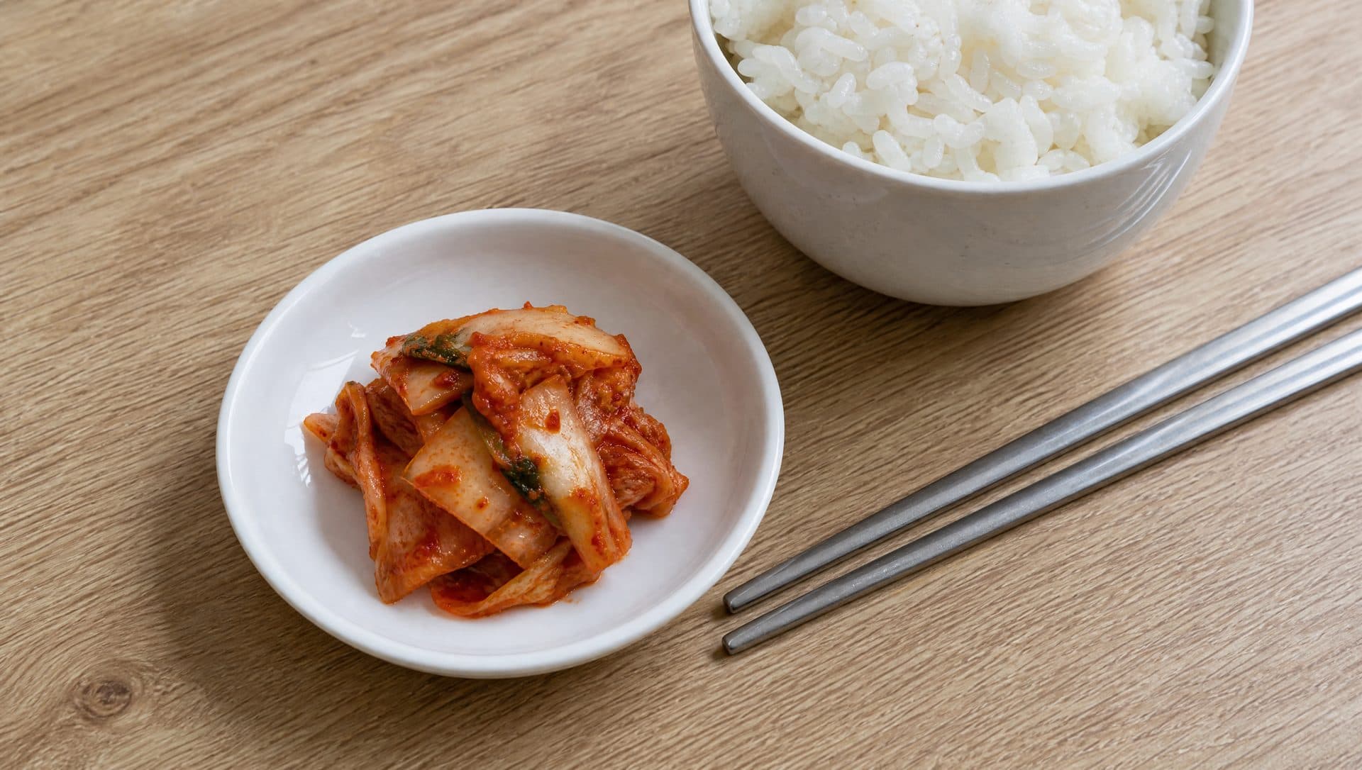 A small white side dish holding a single serving of kimchi beside a bowl of steamed rice and metal chopsticks on a light wooden table