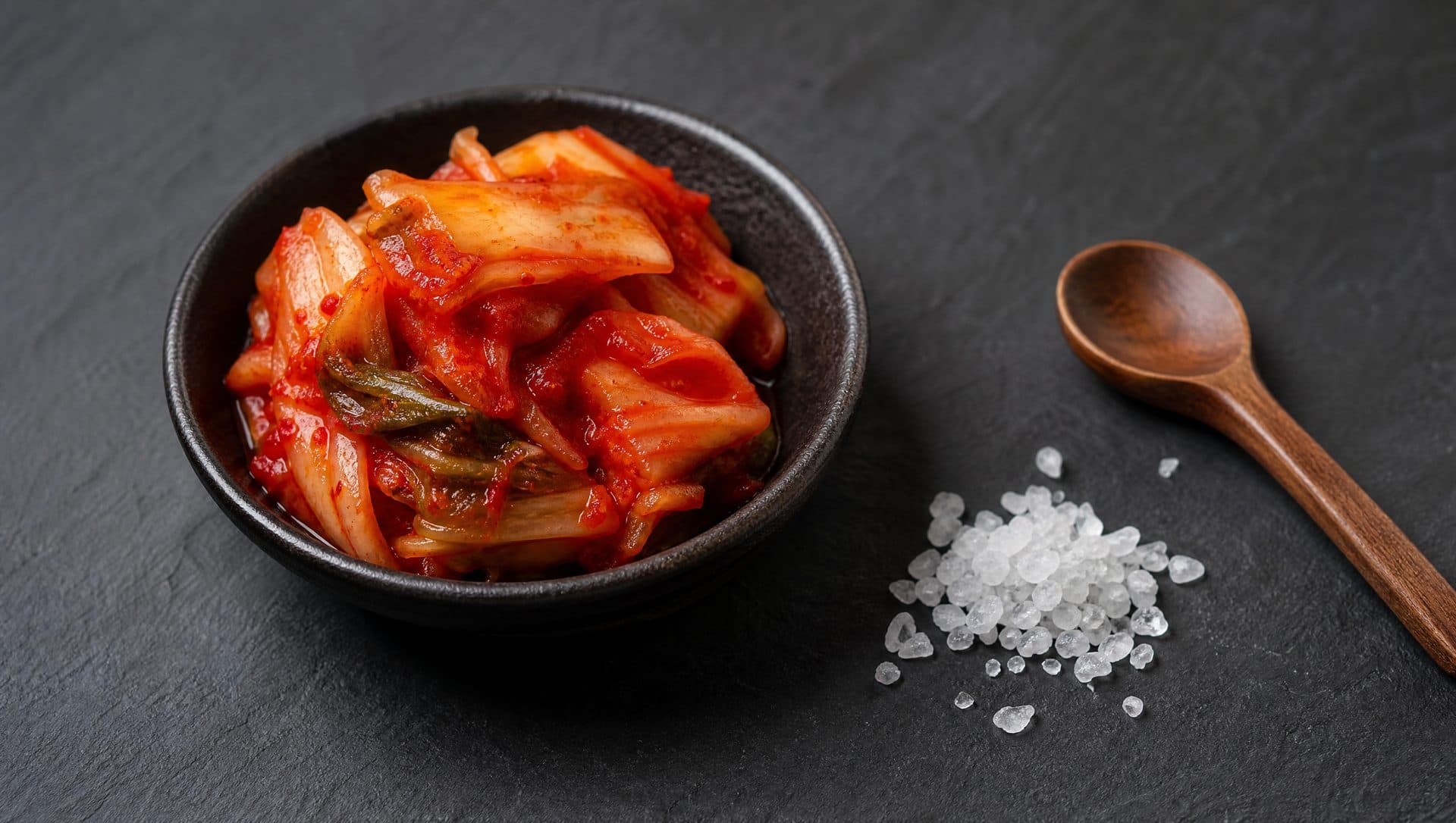 A small dark bowl of kimchi beside a scattering of coarse sea salt and a wooden spoon on dark slate