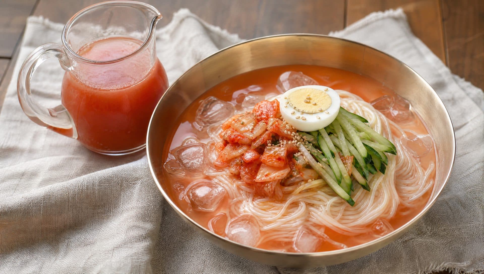 A wide brass bowl of thin white noodles in a pale red iced broth, topped with chopped kimchi, cucumber, half a boiled egg and sesame seeds, next to a small glass jug of strained kimchi brine