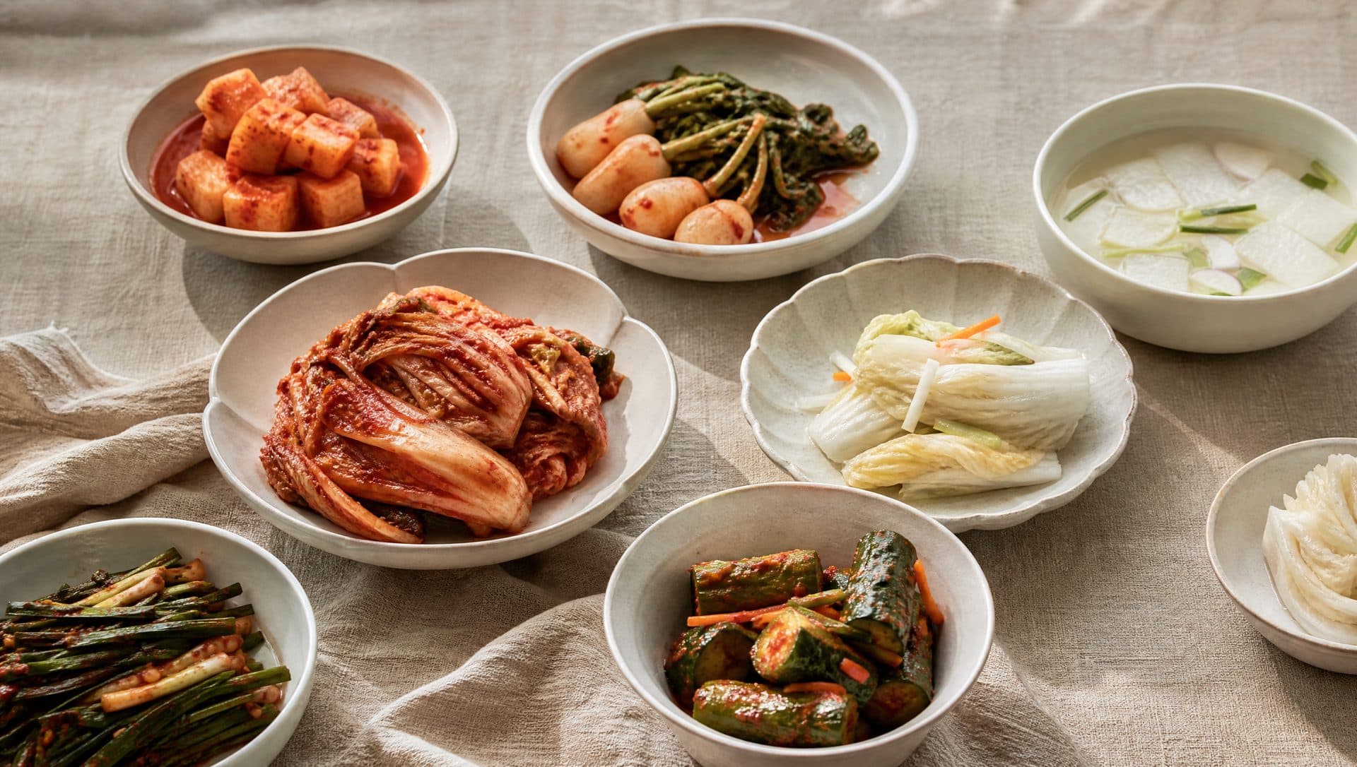 An overhead spread of many kimchi varieties in small ceramic bowls on a linen cloth