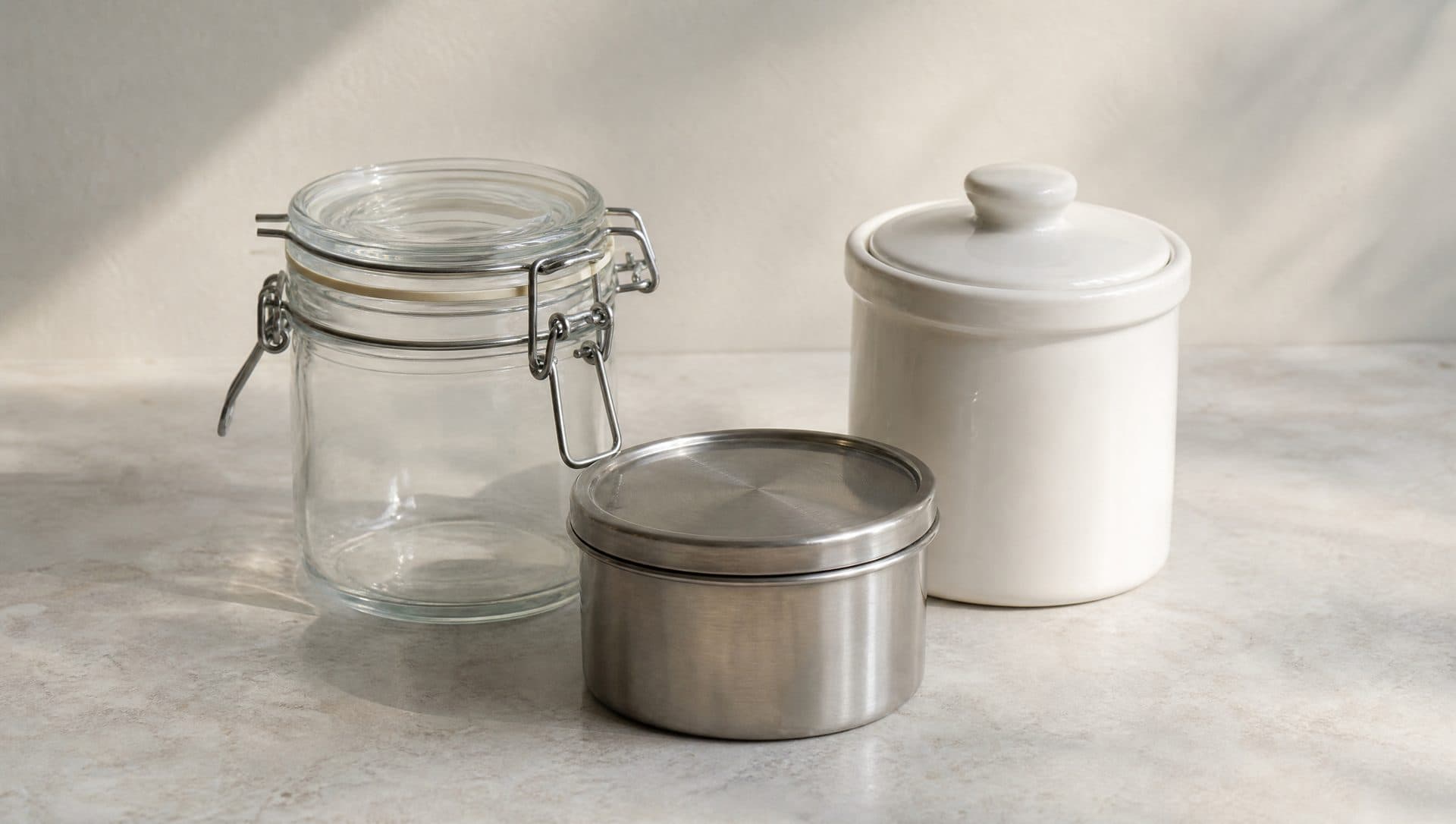 A glass clamp-lid jar, a stainless steel lidded container and a white ceramic crock with a lid on a stone worktop