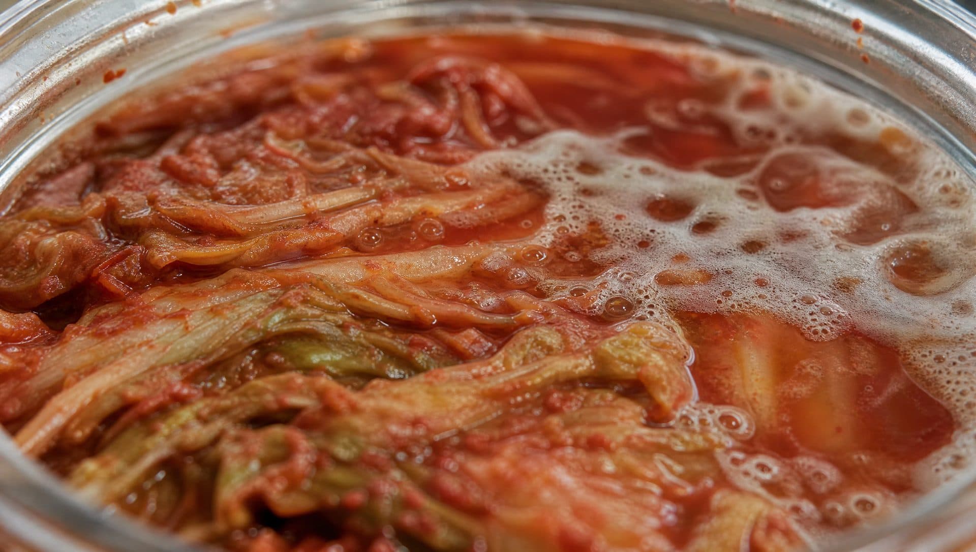 Close-up of the surface of a jar of aged kimchi, leaves pressed under red brine with a faint white film at one edge