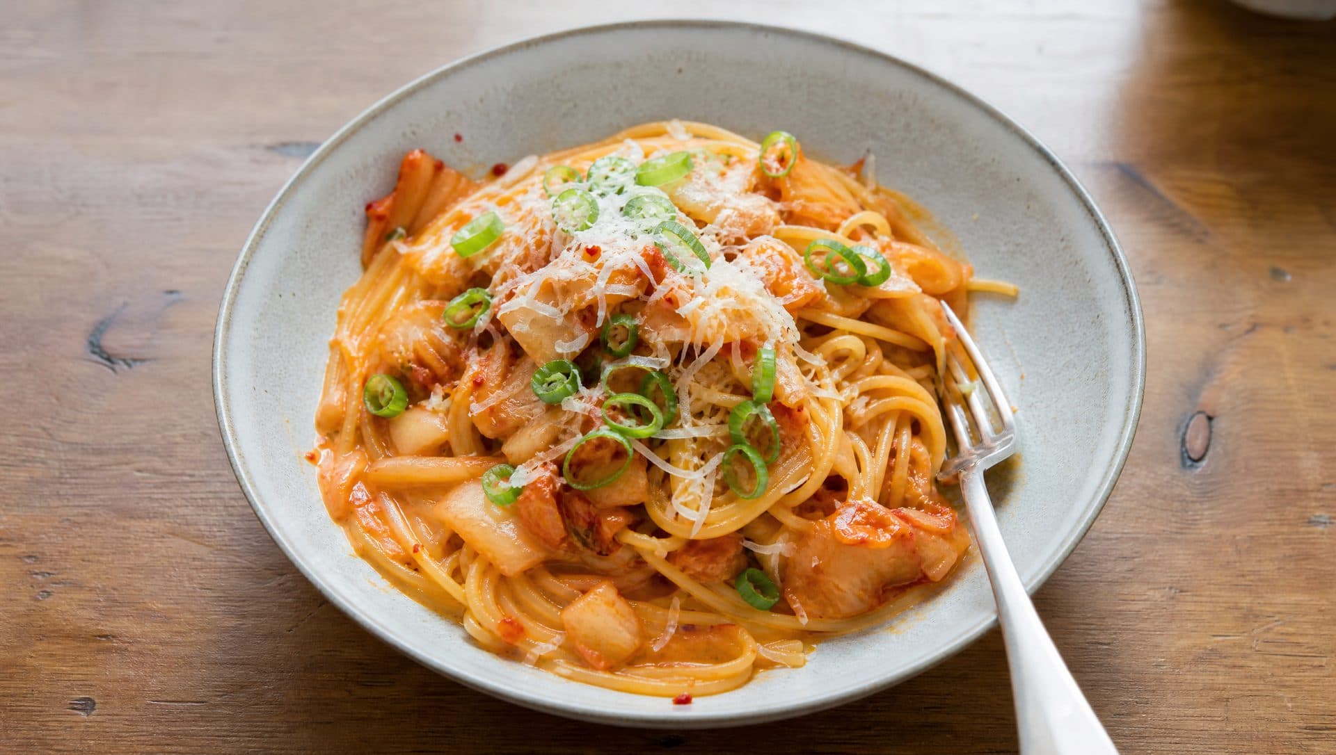 A shallow bowl of spaghetti tossed with chopped red kimchi in a glossy butter sauce, topped with grated cheese and sliced spring onion, on a wooden table in daylight