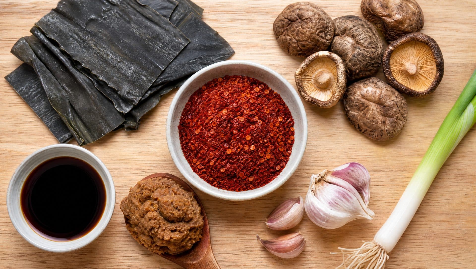 Flat lay on a light wooden board of dried kelp sheets, dried shiitake mushrooms, a small dish of soy sauce and a spoonful of fermented soybean paste around a bowl of red chilli flakes, with garlic and a spring onion