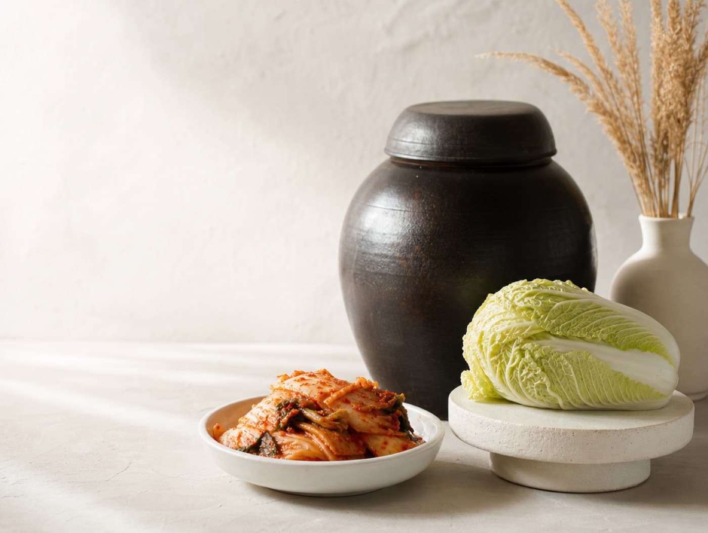 An onggi crock, a whole napa cabbage and a dish of fresh kimchi on a pale surface